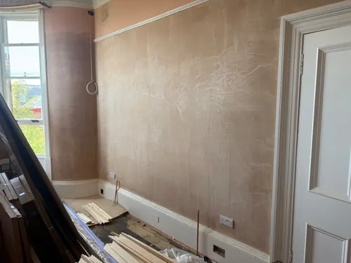 Bay window room stripped to bare plaster with lengths of moulding stacked on the floor